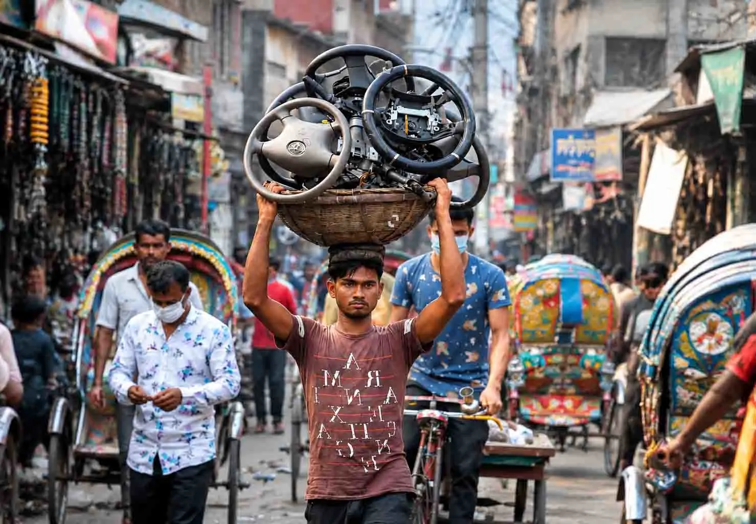 A local street scene in Dholaikhal, Old Dhaka, Bangladesh, featuring a man carrying vehicle parts through a busy lane surrounded by rickshaws, shops, and everyday urban activity.
