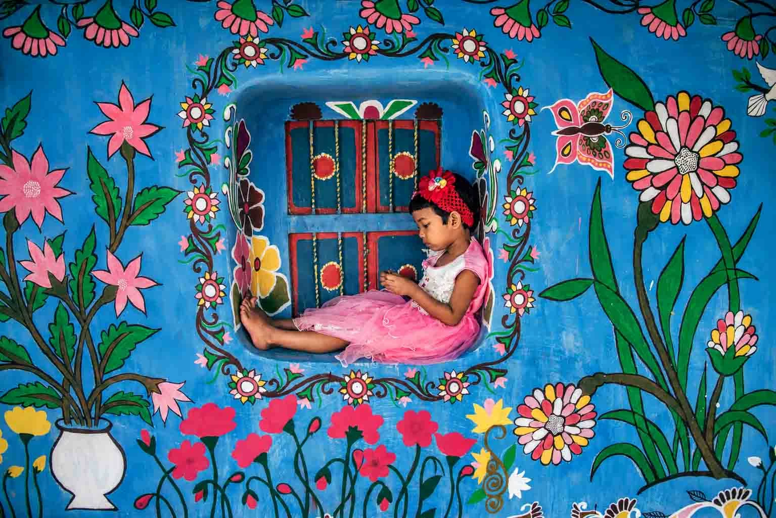 A young girl in a pink dress sitting inside a brightly painted blue window alcove set against a mud house wall adorned with colorful hand-painted floral murals and folk art in Tikoil village. 