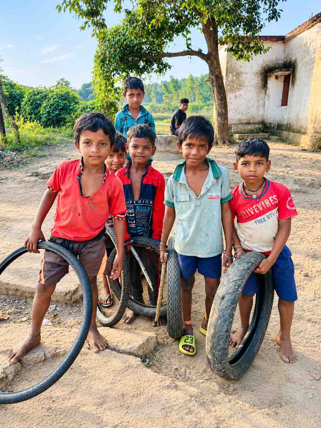 Children playing with old bicycle tires in a rural Bangladeshi village beneath a tree.
