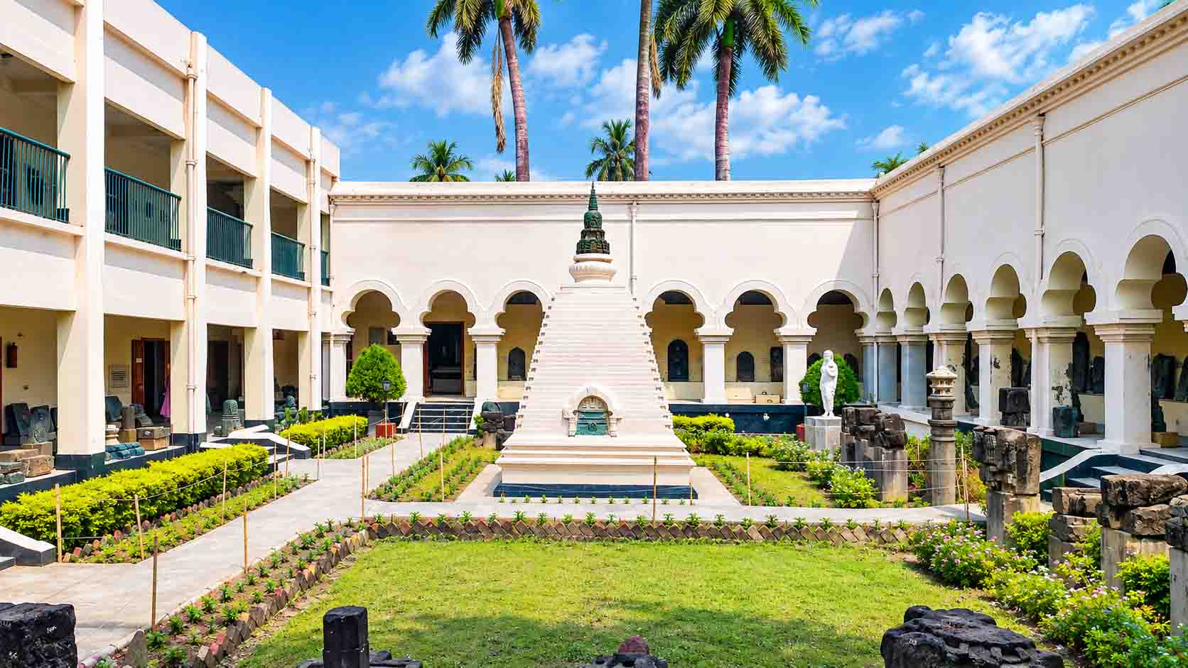 The courtyard of Varendra Research Museum in Rajshahi, Bangladesh, featuring a central white stupa-like monument, arched galleries, ancient sculptures, and landscaped gardens.
