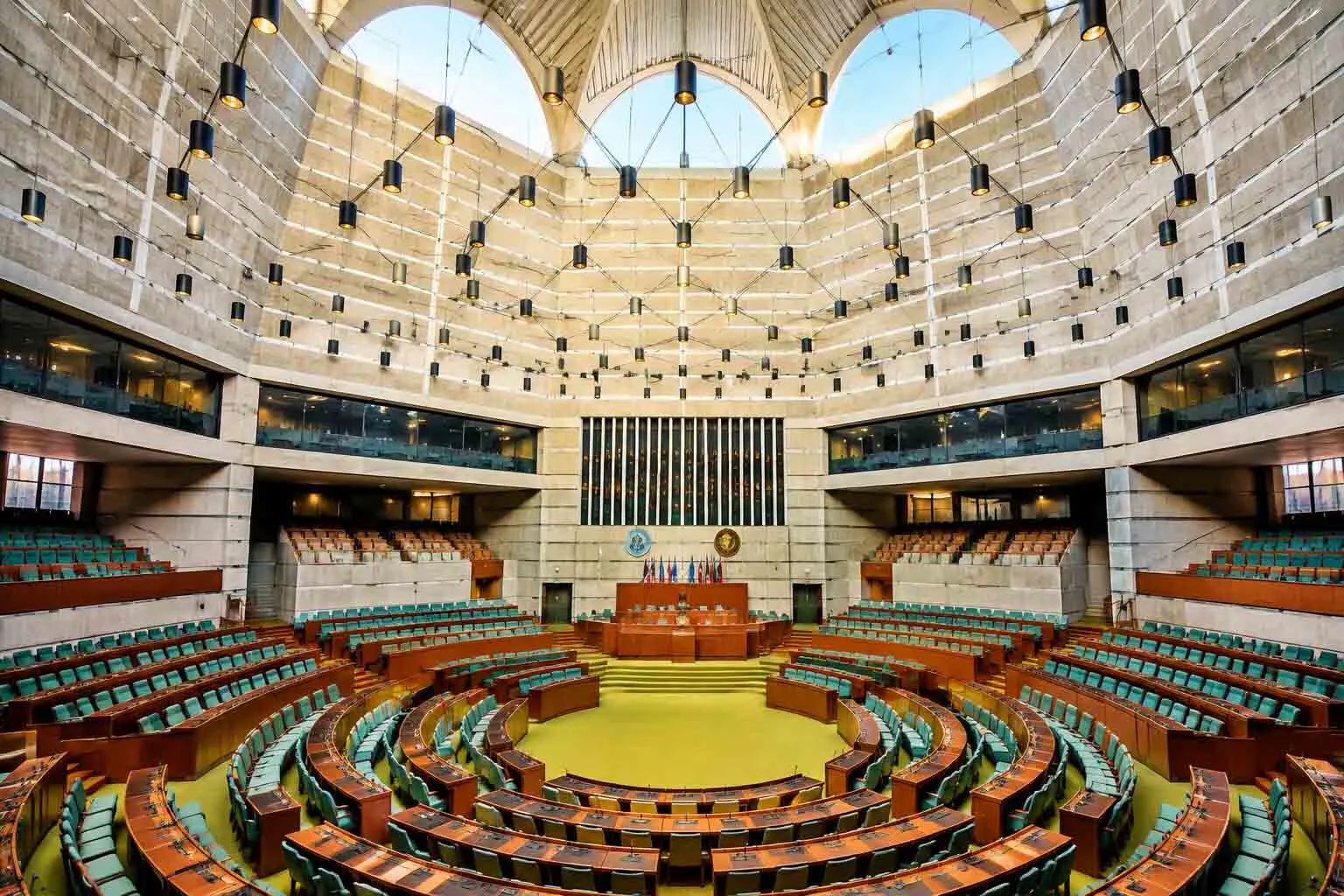 An expansive interior view of the circular parliamentary assembly hall at Jatiya Sangsad Bhaban in Dhaka, featuring green-carpeted floors, concentric curved rows of wooden desks with teal-green chairs, exposed concrete walls, and hanging cylindrical lights under vaulted skylight arches.
