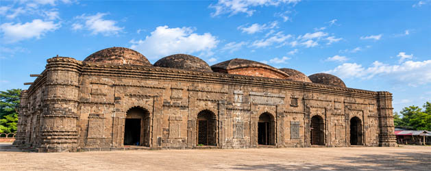 Historic Choto Sona Mosque in Bangladesh featuring intricately carved stone walls, ornate arched entrances, and multiple dark domes beneath a blue sky.