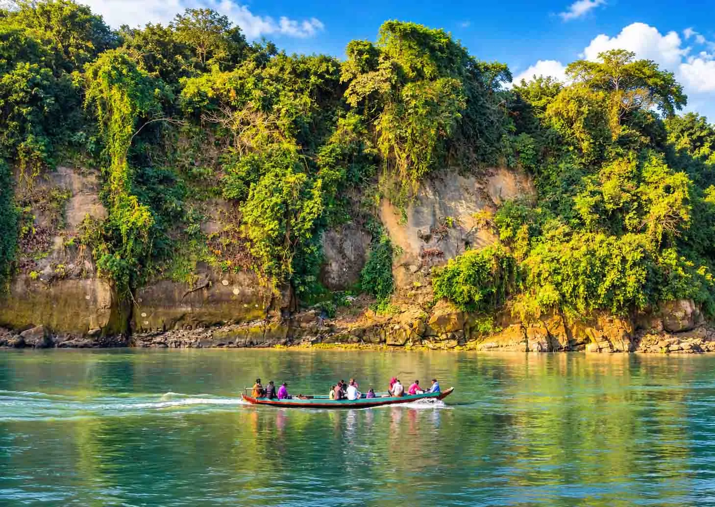 A long wooden passenger boat navigating the clear emerald waters of the Sangu River in Bandarban, backed by steep sandstone cliffs draped in dense green jungle under a bright sky. 