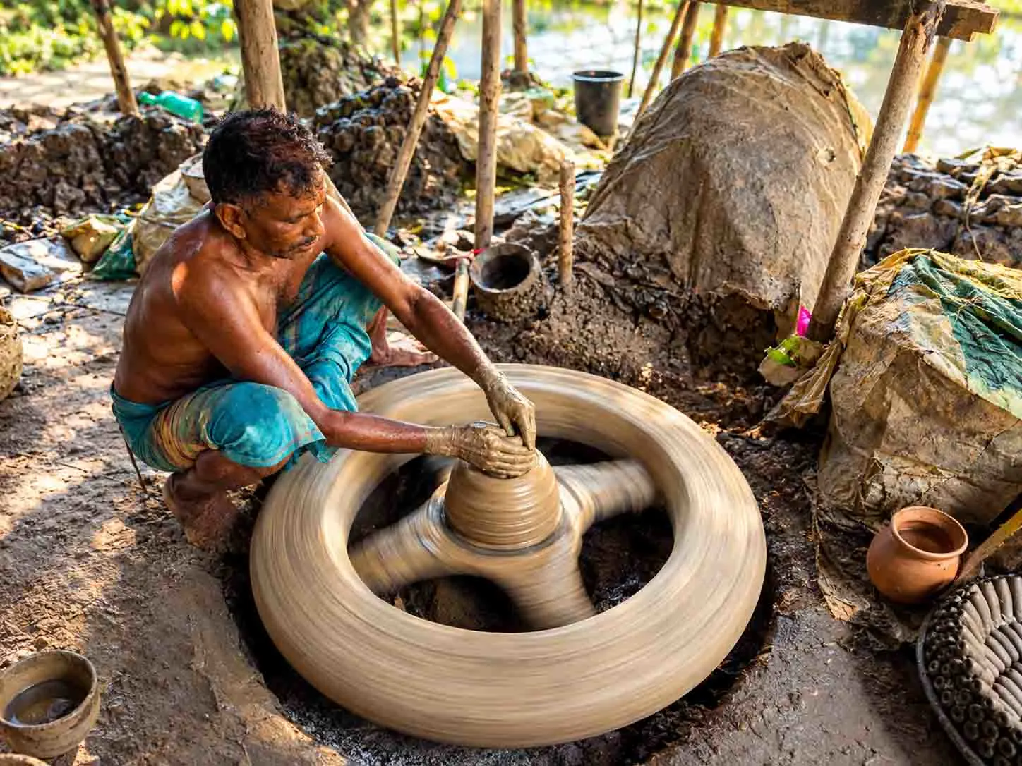 A Bangladeshi potter shaping a clay pot on a traditional spinning pottery wheel in a rustic workshop.