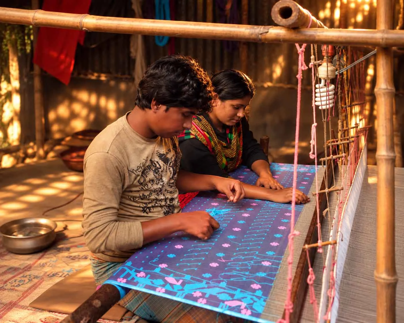 A man and woman artisan sitting side-by-side at a wooden handloom frame, carefully hand-weaving intricate cyan and pink floral motifs onto purple Jamdani fabric using thread needles in a village workshop.