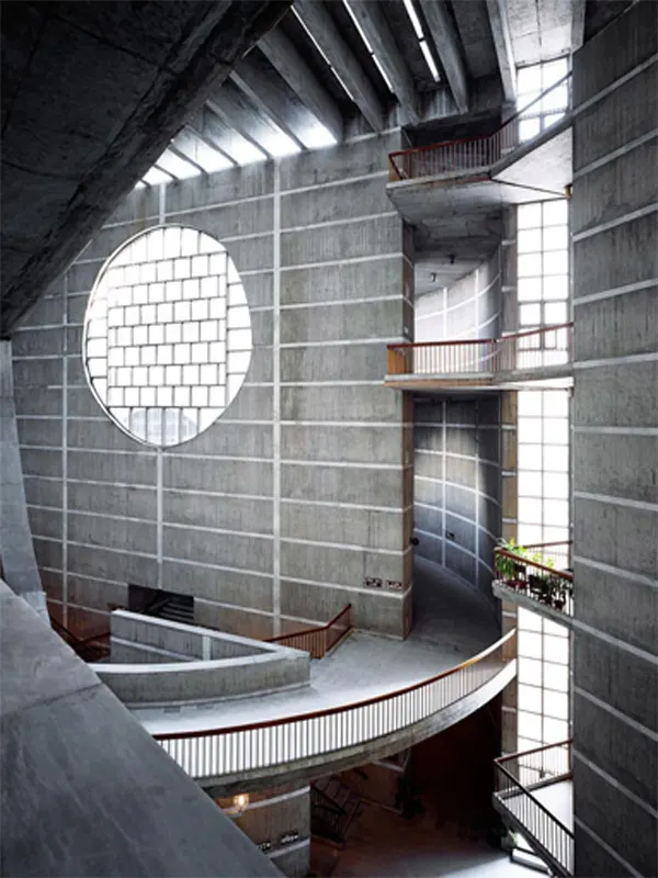 A dramatic vertical interior view of the monumental concrete corridors inside Jatiya Sangsad Bhaban in Dhaka, featuring a large circular cutout window, skylit ceilings, and sweeping curved pedestrian ramps with wooden handrails.