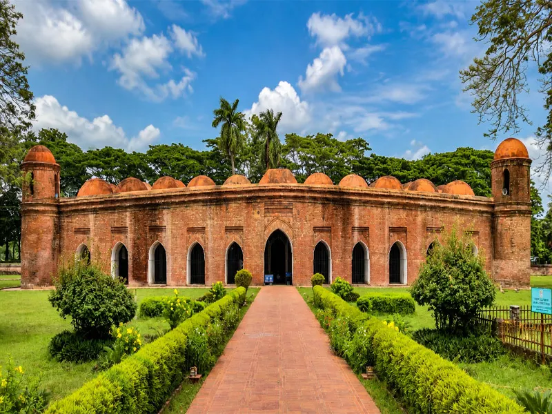 A wide front view of the red-brick Sixty Dome Mosque featuring multiple arched entrances, domed corner towers, and terracotta domes behind a paved brick walkway and manicured green gardens under a blue sky.