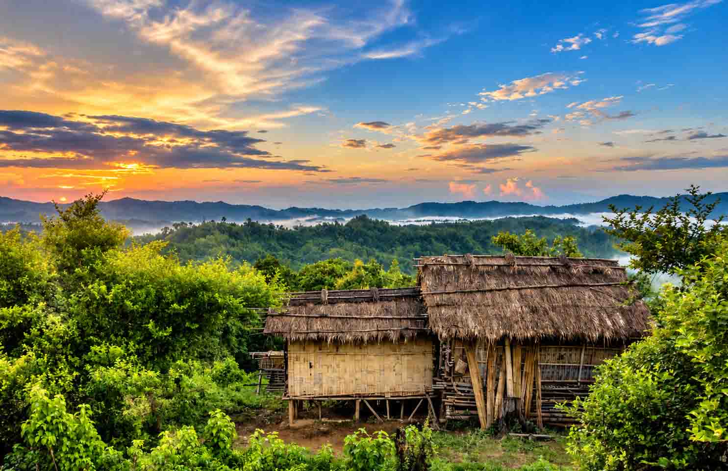 A high-angle shot of a traditional bamboo stilt house with a thatched roof, nestled among lush green foliage in Sajek Valley, looking out over mist-shrouded hill ranges under a golden sunset sky. 