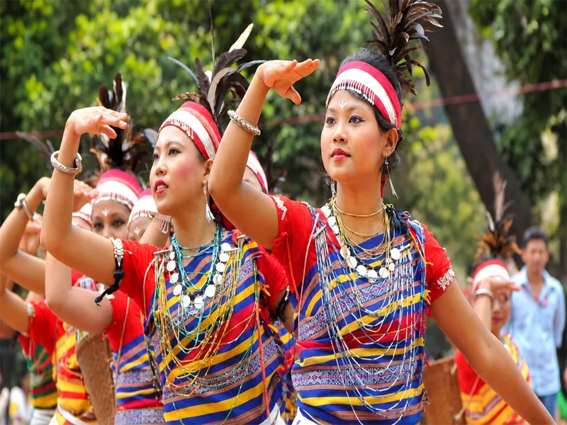 Garo indigenous women dancers wearing feathered headbands, multi-colored striped attire, and layered silver necklaces performing graceful hand gestures during a traditional harvest festival.
