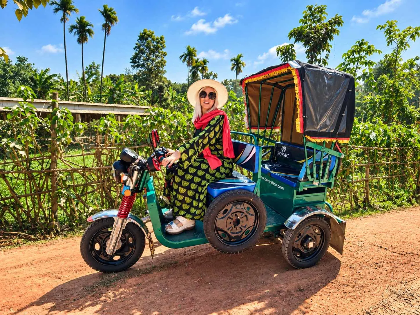 A Romanian female tourist riding a local three-wheeler on a rural road in Bangladesh, surrounded by tropical greenery and village scenery.