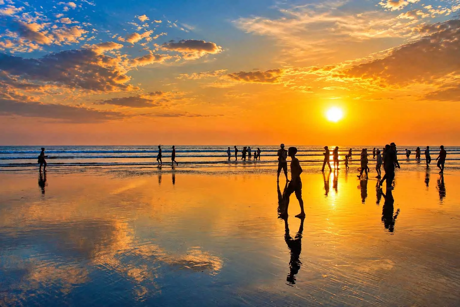 Silhouettes of beachgoers walking along the wet, reflective sandy shore of Cox's Bazar during a brilliant golden hour sunset over the ocean, with glowing yellow-orange skies mirrored in the shallow tide. 