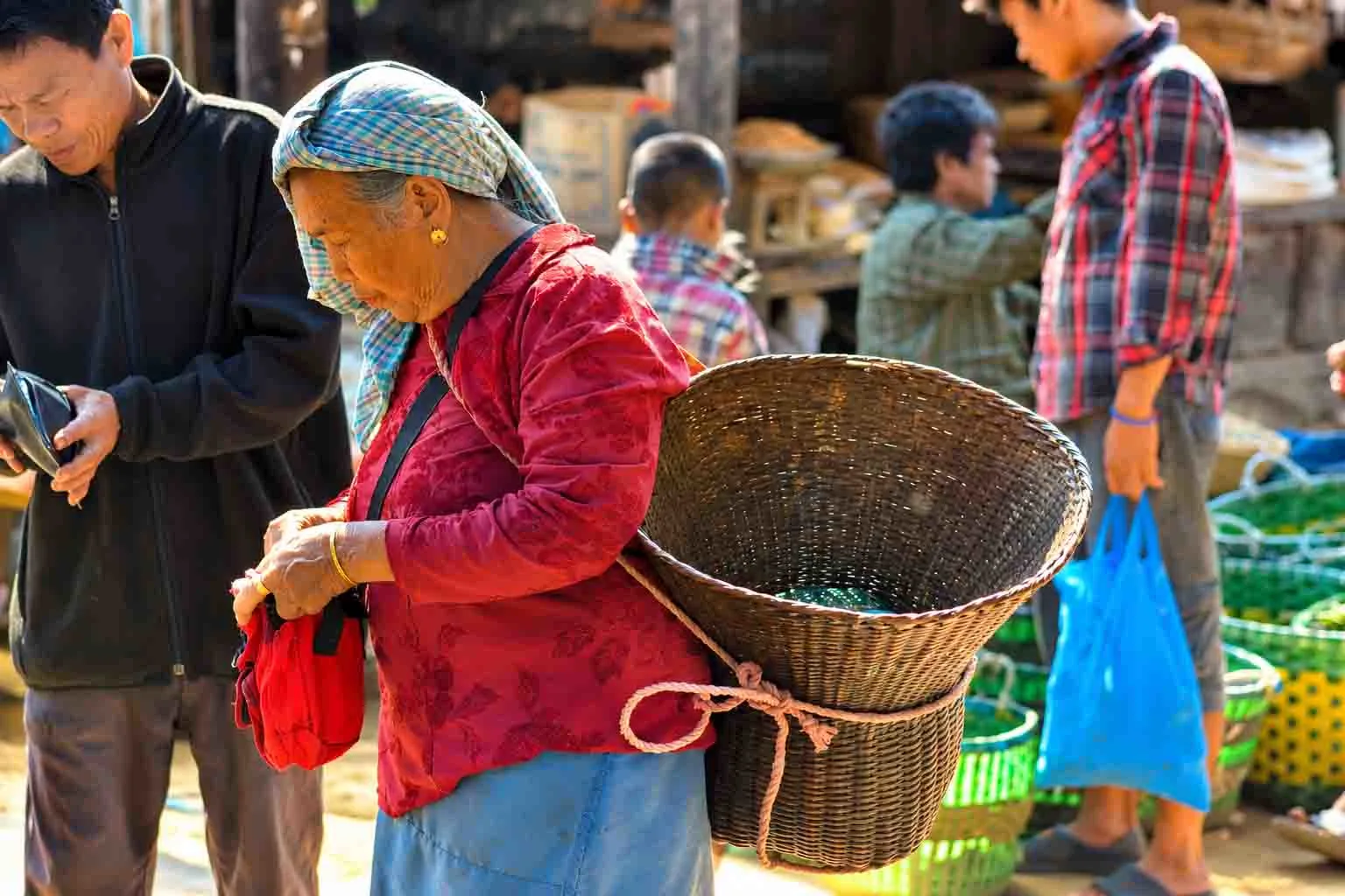 An elderly indigenous woman wearing a red blouse and checkered headscarf carrying a traditional woven bamboo basket on her back in a busy local market in Bandarban.