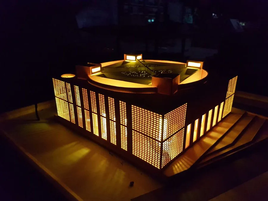An illuminated architectural scale model of the Bait Ur Rouf Mosque in Dhaka, designed by Marina Tabassum, showing its signature square perimeter, inner cylindrical prayer chamber, and glowing perforated brick jali walls.