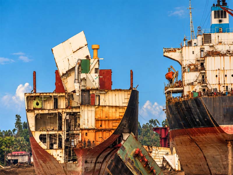 A wide daytime shot of massive ocean vessels being dismantled on the mudflats of Chittagong Ship Breaking Yard, showing a cross-section of a partially cut ship hull against a bright blue sky. 