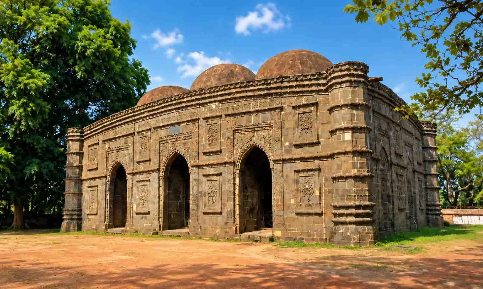An exterior view of the historic dark stone Kusumba Mosque featuring three arched entryways, carved stone relief panels, octagonal corner towers, and stone domes set against green trees and a clear blue sky.