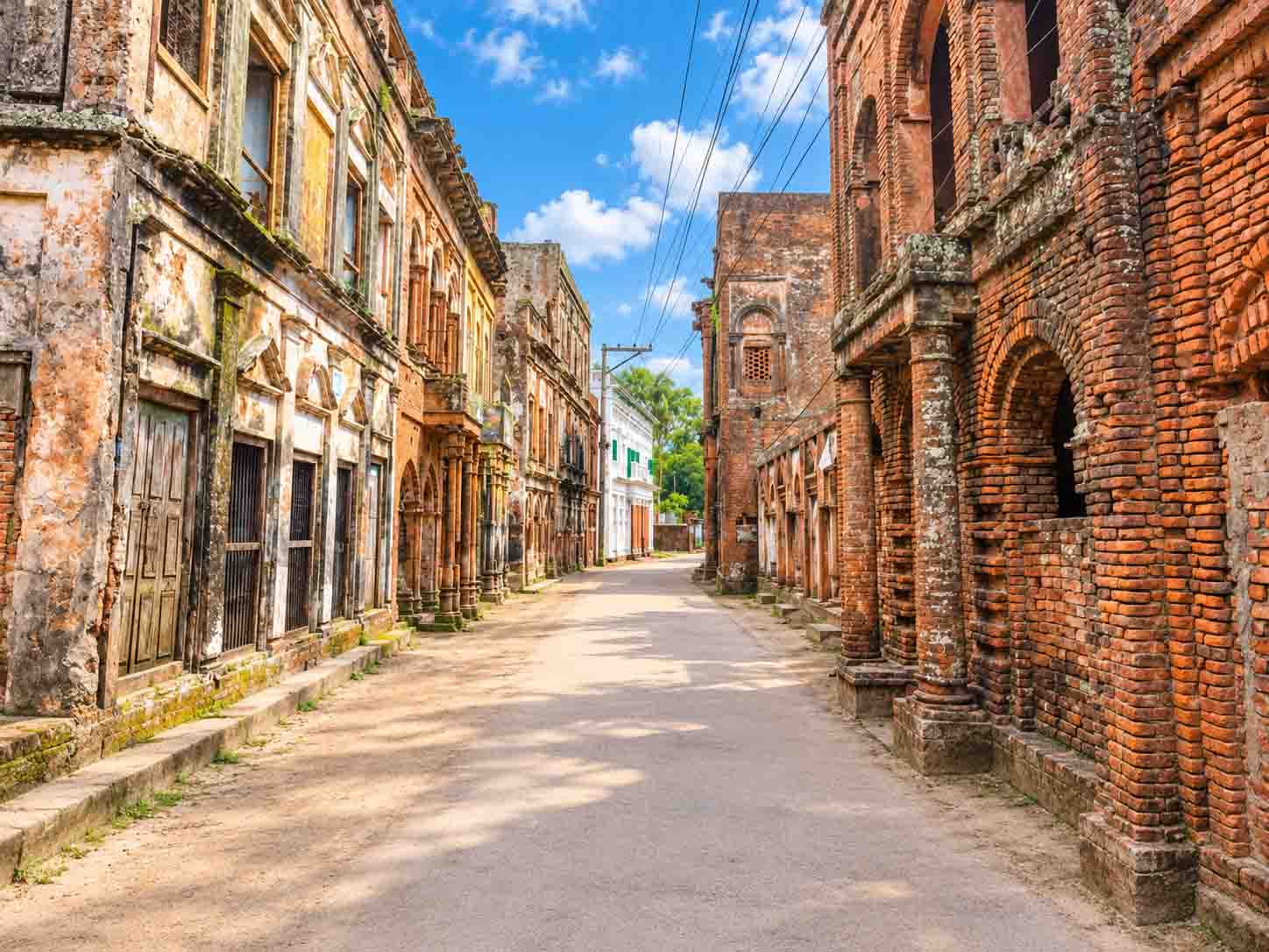 A daytime street view of Panam Nagar in Sonargaon, Bangladesh, featuring historic colonial-era red brick buildings with weathered facades and intricate archways lining both sides of a paved road under a blue sky. 