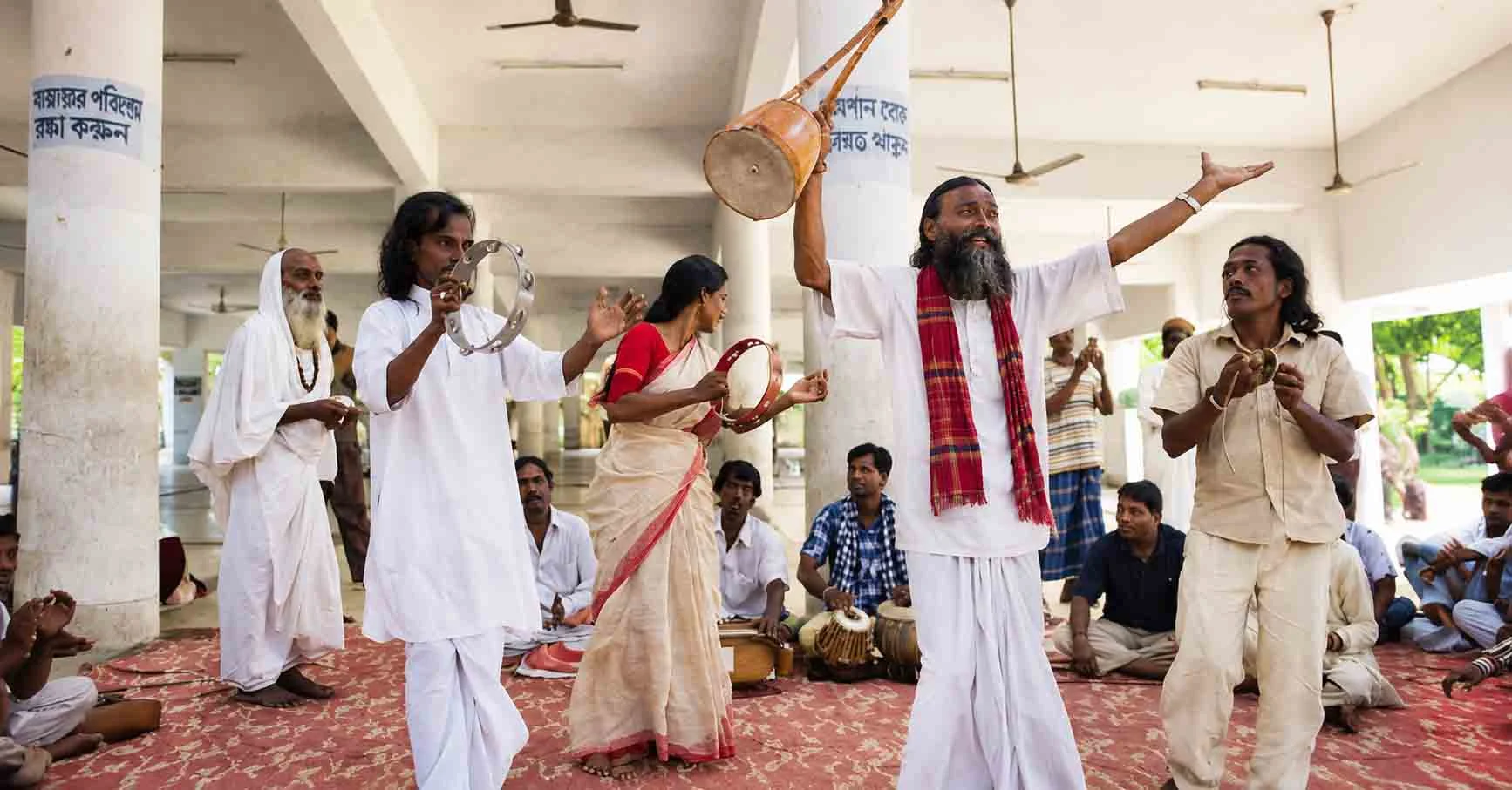 A group of Baul musicians in traditional white attire performing folk music with a raised Ektara, Dubki tambourine, and Mandira cymbals inside the open pillar pavilion at Lalon Akhra in Kushtia. 