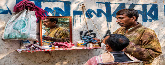 A traditional street barber provides a shave to a seated customer at an outdoor roadside setup in Dhaka, Bangladesh, with a mirror, barber tools, and a weathered wall featuring Bengali writing in the background.