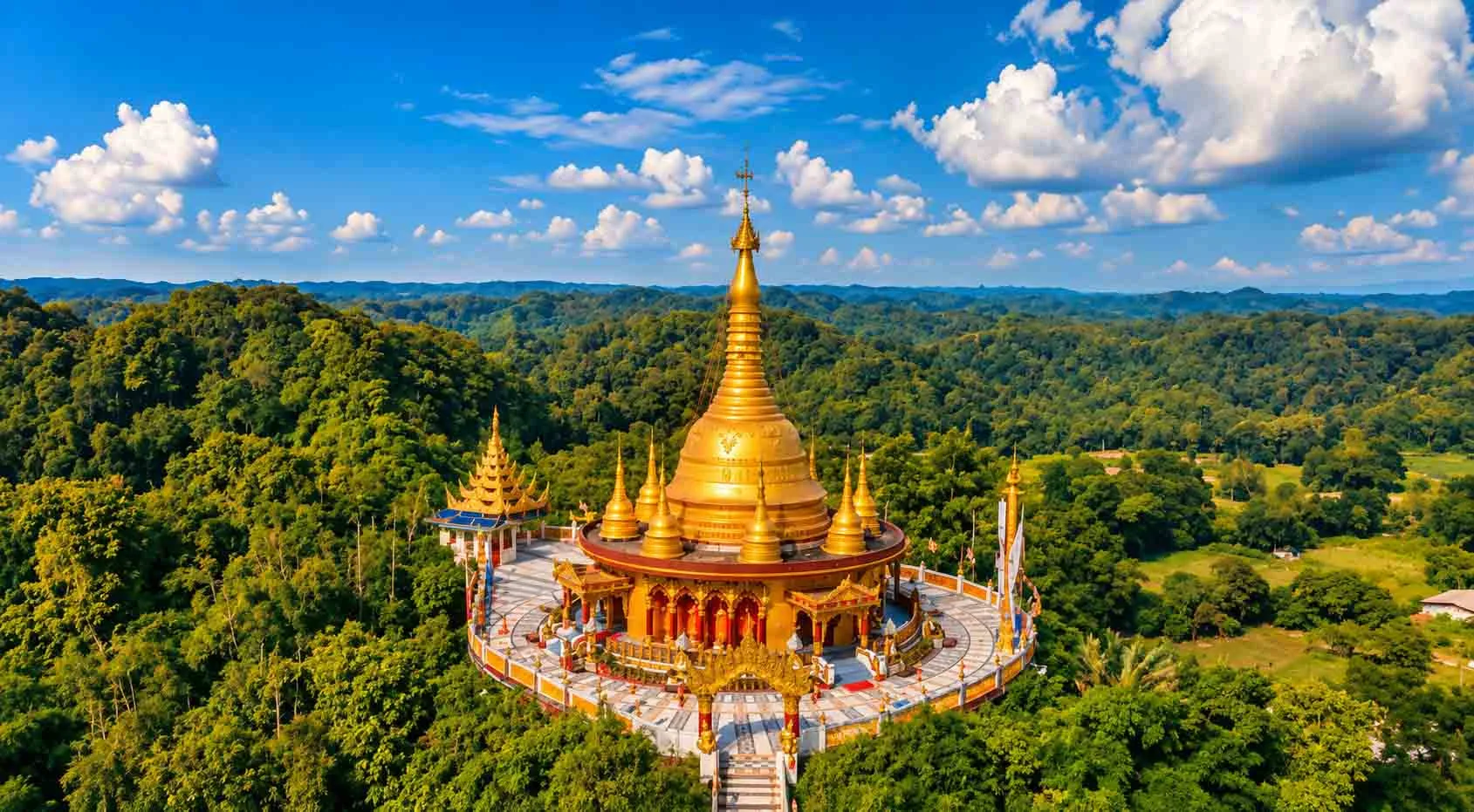 An aerial high-angle shot of the golden Buddha Dhatu Jadi pagoda perched atop lush green tropical hills in Bandarban under a vibrant blue sky with fluffy white clouds. 