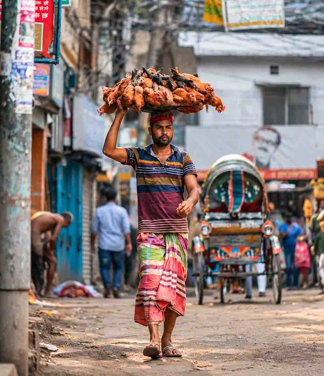 A street vendor carrying a basket of chickens on his head while walking through a busy lane in Dhaka, Bangladesh, with a traditional rickshaw and local shops in the background.
